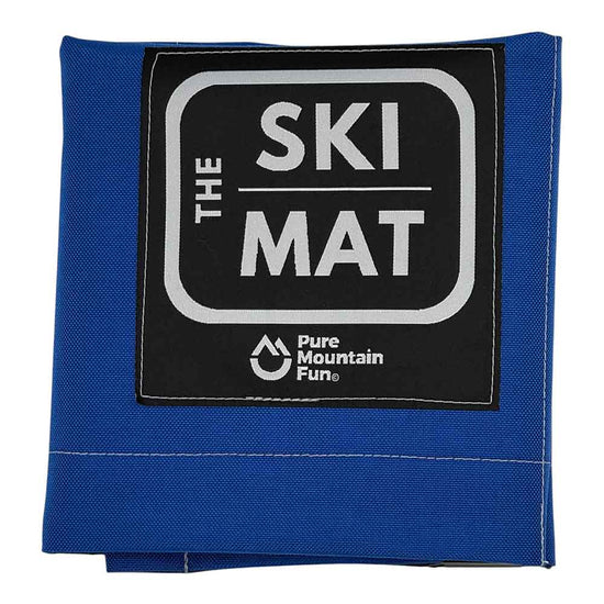 A folded blue ski mat with a black square label on the front that reads 'THE SKI MAT' and shows the 'Pure Mountain Fun' logo, photographed on a white background.