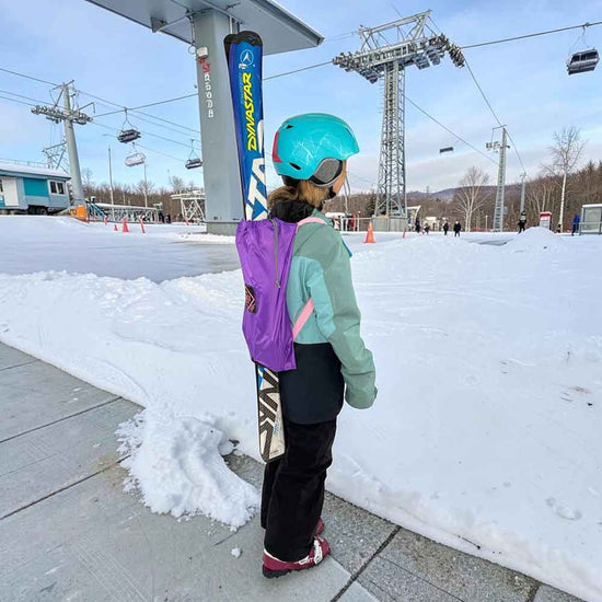 A child standing on a snowy ski resort, viewed from behind, wearing a mint green jacket, black pants, a turquoise helmet, and wearing a purple ski backpack with a ski attached. Ski resort towers and chairlifts are visible in the background.