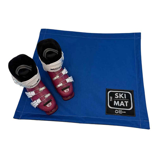 A blue ski mat with a black 'The Ski Mat' logo patch in the bottom right corner, shown on a white background with a pair of ski boots placed on top.